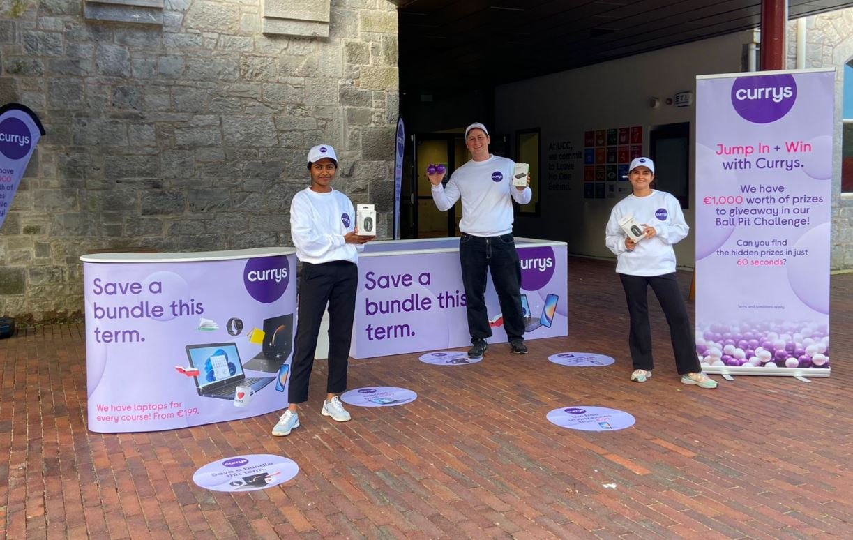 The Currys student campaign stand on a campus concourse, three staff in branded caps.
