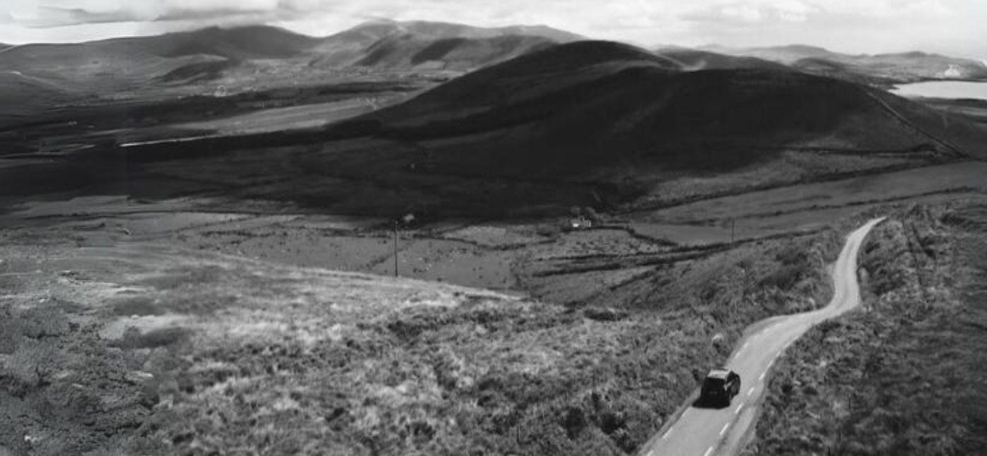 A single car on a road through open hills, in black and white.