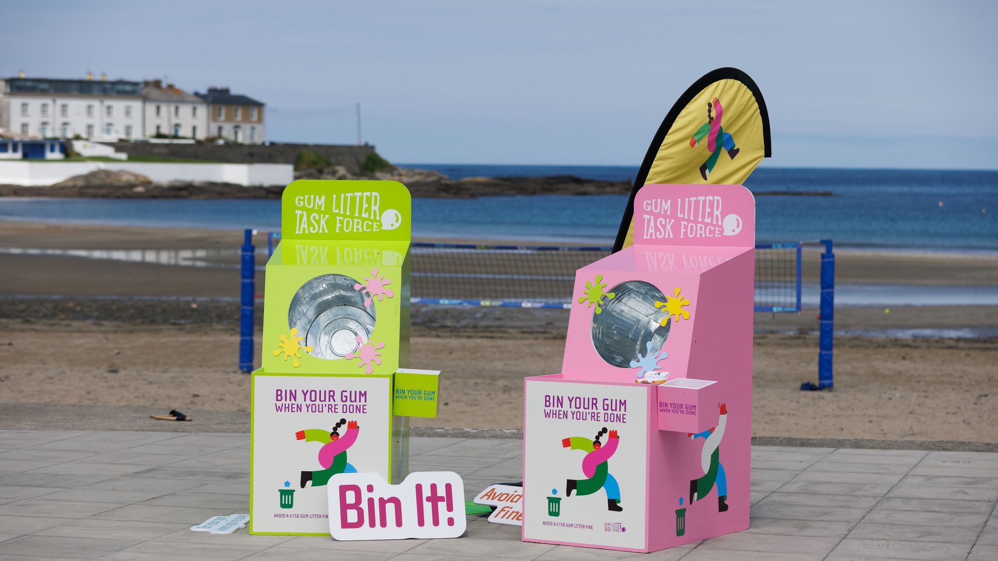 Two branded Gum Litter Taskforce units on a seafront promenade, reading Bin your gum when you are done, with the beach behind them.