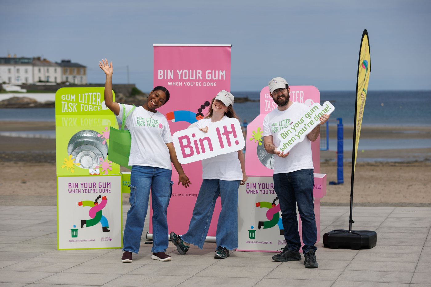 Three brand ambassadors waving beside the Gum Litter Taskforce units on a seafront promenade.